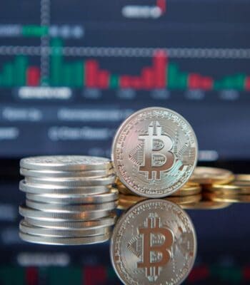 Abgebildet sind mehrere Münzen, darunter eine Bitcoin-Münze, vor einem Hintergrund mit Finanzdiagrammen. Die Darstellung symbolisiert die Relevanz von Kryptowährungen und Blockchain-Technologie im aktuellen Finanzumfeld.