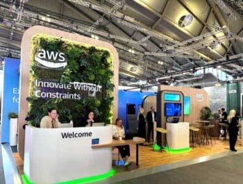 Der Stand von AWS auf der SIBOS 2025 präsentiert sich modern und einladend. Begrüßungstresen und interaktive Displays: Die Gestaltung mit Pflanzen und dem Slogan „Innovate Without Constraints“ soll Innovtion vermitteln.