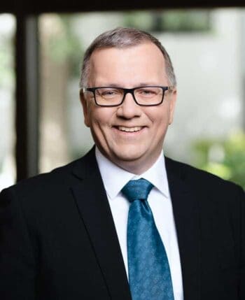 Dirk Strauch in einem schwarzen Anzug mit blauer Krawatte lächelt freundlich. Der Hintergrund ist unscharf und zeigt eine helle, grüne Umgebung. Excel wird häufig zur Verwaltung digitaler Zertifikate verwendet, was jedoch ineffizient und riskant ist.