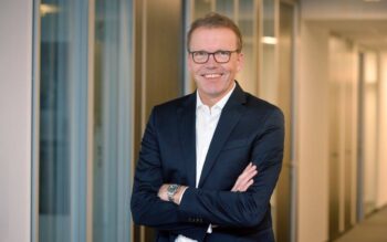 Dominik Schlarmann, stellv. Vorstandsvorsitzender der Sparda-Bank West eG, steht in einem modernen Büroflur. Er trägt einen dunklen Anzug und lächelt, während er die Arme vor der Brust verschränkt.