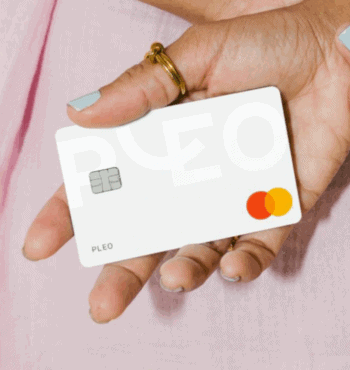 Frauenhand mit goldenem Ring am Daumen und hellblauen Nägeln hält Pleo Mastercard vor pinken Hintergrund in die Kamera.