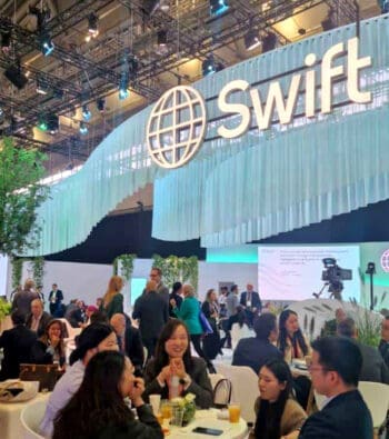 Auf der Sibos (der Hausmesse der SWIFT) zeigt eine lebhafte Interaktion zwischen Teilnehmern. Die Umgebung ist modern gestaltet, mit Pflanzen und einem großen Logo. Diskussionen über Stablecoins, CBDC und Künstliche Intelligenz sind offensichtlich.