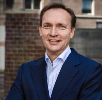 Željko Kaurin, künftig Vorstand für „Digital Banking“ der Atruvia, präsentiert sich in einem formellen Outfit. Der Hintergrund zeigt eine moderne Architektur, die Professionalität und Fortschrittlichkeit symbolisiert.