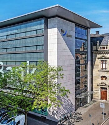Die Abbildung zeigt die moderne Fassade der BBBank in einem urbanen Umfeld. Die Architektur kombiniert Glas- und Steinelemente. Im Vordergrund sind Bäume und Parkplätze sichtbar, was zur Gestaltung des Außenbereichs beiträgt.