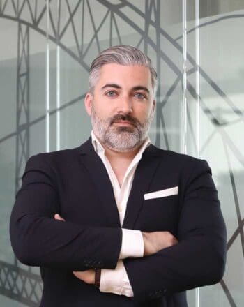 Ein Mann mit grauem Bart und Haar, gekleidet in einen dunklen Anzug, steht mit verschränkten Armen vor einer modernen Glaswand. Die Umgebung vermittelt Professionalität, was die Bedeutung von Cycode für die IT-Sicherheit im Finanzsektor unterstreicht.