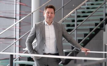 Jens Heilmann, Experte für digitale Transformation, steht an einer Treppe in einem modernen Gebäude. Sein Fokus liegt auf der DORA-Compliance, die für die IT-Migration und den Einsatz von Künstlicher Intelligenz von Bedeutung ist.
