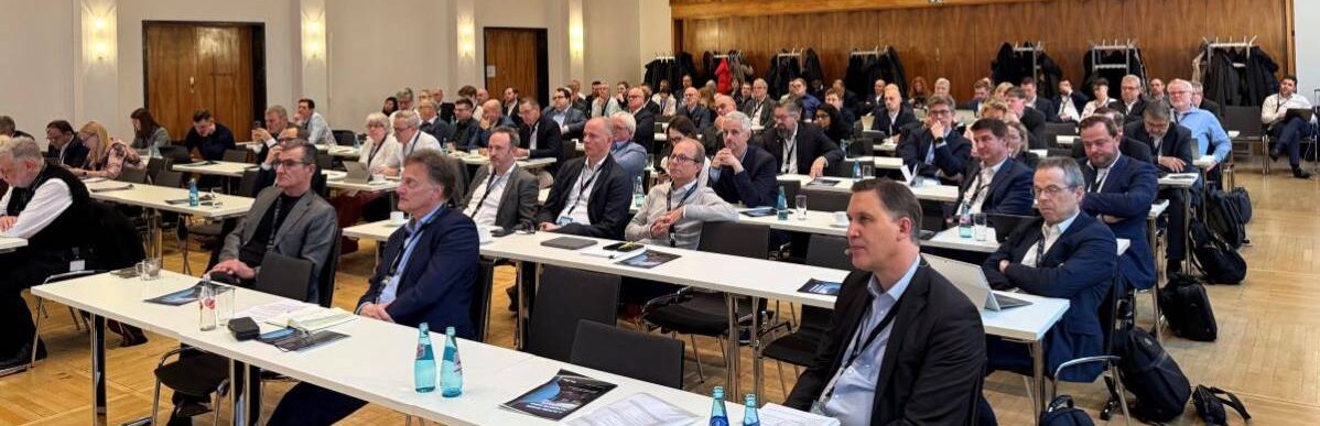 Das Zahlungsverkehrsforum versammelt zahlreiche Fachleute in einem Konferenzraum. Die Teilnehmer sind aufmerksam und konzentriert, während sie den Präsentationen folgen. Auf den Tischen sind Wasserflaschen und Unterlagen platziert, die zur Veranstaltung gehören.