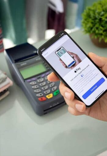 Ein Handheld-Gerät zeigt eine Zahlungsanwendung auf einem iPhone, während eine Hand das Gerät hält. Im Hintergrund ist ein Kartenlesegerät sichtbar. Die Darstellung verdeutlicht die Nutzung von NFC-Zahlungen ohne Apple Pay.