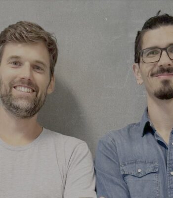 Zwei Männer stehen nebeneinander vor einer grauen Wand. Der linke Mann trägt ein graues T-Shirt, der rechte ein blaues Hemd. Beide lächeln. Diese Zusammenarbeit zwischen CONDA und StartMatch zielt darauf ab, die Emissionen durch KI zu vereinfachen