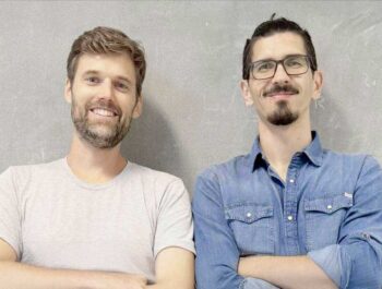 Robert Kopka und Oliver Lukesch, Gründer von StartMatch, stehen nebeneinander vor einer grauen Wand. Beide präsentieren ein freundliches Lächeln, was die positive Ausstrahlung ihrer Zusammenarbeit im Kontext von Conda unterstreicht.