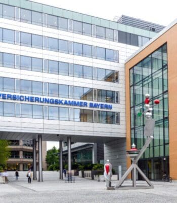 Die Fassade der Versicherungskammer Bayern ist modern gestaltet, mit großen Fenstern und einer auffälligen Beschriftung. Vor dem Gebäude befindet sich eine künstlerische Skulptur. Mehrere Personen bewegen sich im Eingangsbereich.