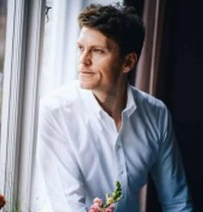 Ein Mann in einem weißen Hemd sitzt am Fenster und blickt nachdenklich nach außen. In der Nähe befinden sich Blumen. Der Kontext verweist auf die Rolle von Pleo im Finanzmanagement.