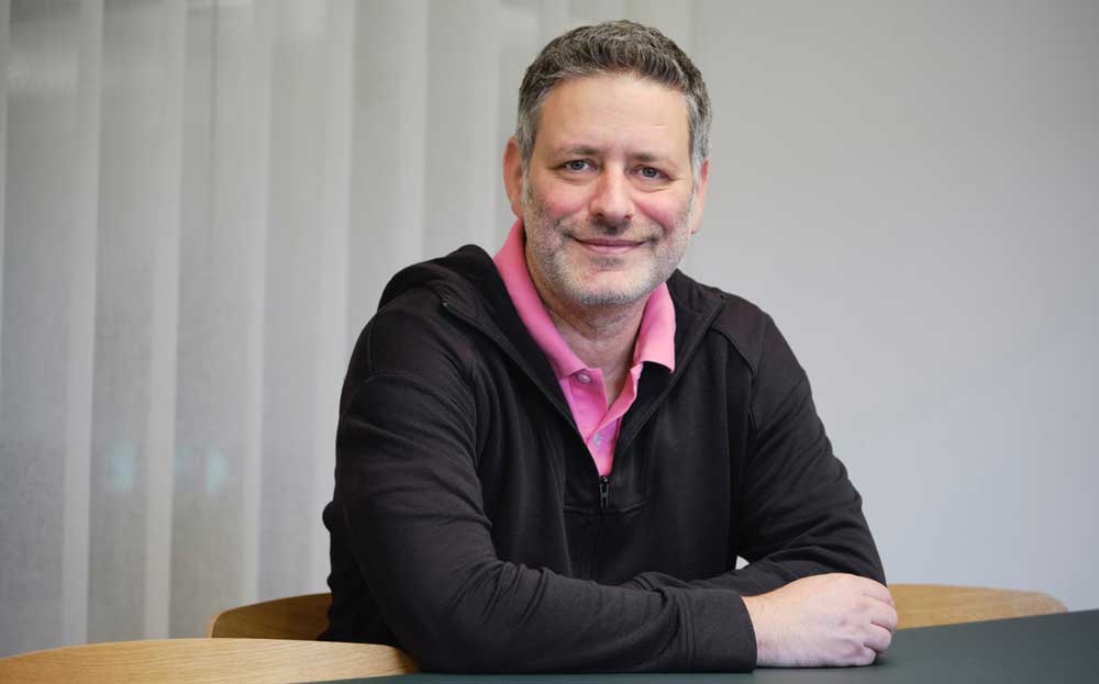 Ein Mann mit grauen Haaren und einem freundlichen Lächeln sitzt an einem Tisch. Er trägt ein schwarzes Oberteil und ein pinkes Hemd. Der Hintergrund ist hell und neutral. Im Kontext des Interviews wird UnzerAI als interner KI-Assistent thematisiert.