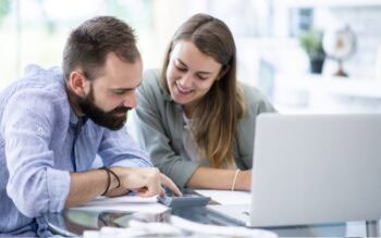 Mann und Frau sitzen an einem Tisch wir einem Laptop und beugen sich über einen Taschenrechner. Worldline Grafik zur PSA Meldung.