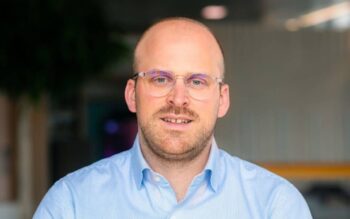 Arne Rötzmeier, Leiter Value Added Services, Visa Zentraleuropa, ist in einem modernen Büro abgebildet. Er trägt ein hellblaues Hemd und eine Brille. Der Hintergrund zeigt unscharfe Elemente, die auf eine professionelle Umgebung hinweisen.