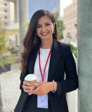 Aynur Colpan, Referentin in der Niedersächsischen Staatskanzlei, steht in formeller Kleidung mit einem Kaffeebecher in der Hand. Der Hintergrund zeigt eine städtische Umgebung, die auf eine Veranstaltung hinweist. Das Bild thematisiert die Bedeutung von Payment