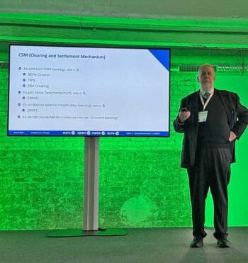 Hans-Rainer van den Berg (CEO euro-V) präsentiert auf der FinTra World in Köln Informationen zur Clearing- und Abwicklungsmechanik (CSM). Auf dem Bildschirm sind relevante Punkte zu sehen, die die Notwendigkeit externer Infrastruktur thematisieren.