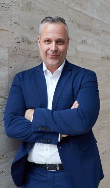 Torsten Krieger, CEO der Reisebank, präsentiert sich in einem formellen Outfit. Der Hintergrund aus Stein vermittelt Professionalität. In der Diskussion um KI-Agenten und deren Einfluss auf das Finanzwesen spielt er eine zentrale Rolle.