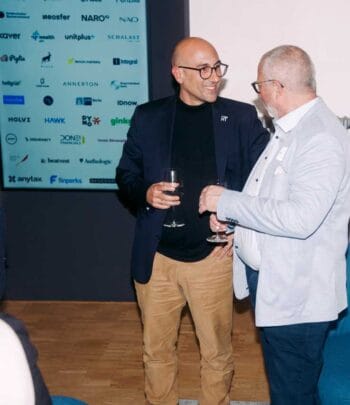 Sebastian Schäfer, Geschäftsführer HoFT, unterhält sich mit einem weiteren Teilnehmer bei der Eröffnungsfeier des House of Finance and Tech in Berlin. Im Hintergrund sind Logos verschiedener FinTech-Unternehmen sichtbar, die das Event unterstützen.