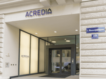 Die Eingangstür zur Acredia Versicherung ist sichtbar, mit dem Unternehmenslogo prominent über dem Eingang. Die Fassade zeigt moderne architektonische Elemente und eine klare Beschilderung der Adresse in der Himmelpfortgasse.