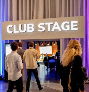 Der CLUB STAGE des FIBE 2026 in Berlin präsentiert eine dynamische Eventstruktur. Teilnehmer bewegen sich in Richtung der Bühne, wo interaktive Diskussionen stattfinden. Die Atmosphäre ist geprägt von Festival- und Club-Elementen.