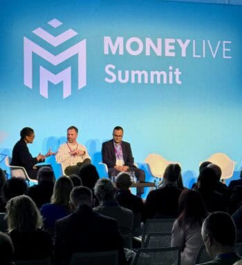 Auf dem MoneyLIVE Summit diskutieren mehrere Experten auf einer Bühne. Im Vordergrund sind drei Personen zu sehen, die miteinander sprechen. Der Hintergrund ist in einem hellen Blau gehalten, mit dem Logo der Veranstaltung prominent platziert.