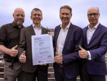 Die Zertifikatsübergabe habe am 13. März im Hamburger Sitz von Laiqon stattgefunden. Vier Personen halten das ISO 27001-Zertifikat und zeigen Daumen hoch. Die Atmosphäre ist feierlich und zeigt den Erfolg der Zertifizierung im Bereich Datenschutz.