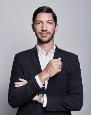 Ein Mann in einem schwarzen Anzug mit weißem Hemd präsentiert sich selbstbewusst. Seine Arme sind vor der Brust verschränkt, während er leicht lächelt. Diese Darstellung könnte im Kontext der Vision Chain stehen, die Finanzinstitute mit Blockchain-Technologie verbindet.