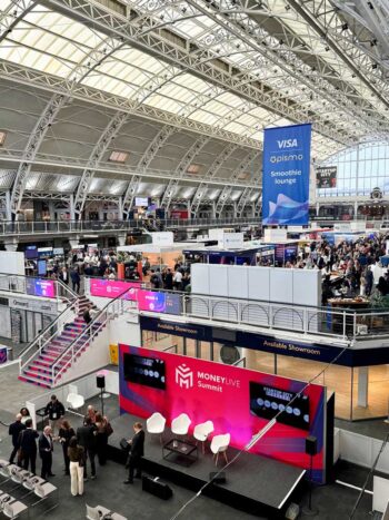 Der MoneyLive Summit findet in einer großen Messehalle statt, in der zahlreiche Teilnehmer an Ständen und Diskussionen teilnehmen. Eine Bühne mit Sitzgelegenheiten ist im Vordergrund sichtbar, während im Hintergrund Banner und Stände von verschiedenen Unternehmen präsentiert werden.