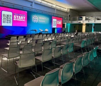 Ein Veranstaltungsraum mit mehreren Reihen von Stühlen, die auf eine Bühne ausgerichtet sind. Im Hintergrund sind große Bildschirme mit QR-Codes und der Aufschrift „START NETWORKING“ sowie dem Logo der MoneyLIVE Summit zu sehen. Das Stilleben Stühle ...