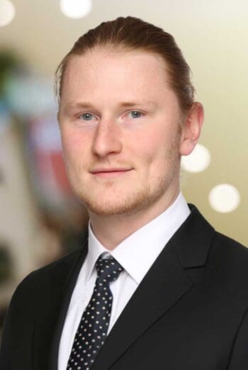 Tom Schmelzer, Assistant Manager bei KPMG, ist in formeller Kleidung abgebildet. Der Hintergrund ist unscharf und zeigt helle Lichter, die eine professionelle Atmosphäre schaffen. Das Bild thematisiert die Relevanz von Instant Payments im Bankensektor.