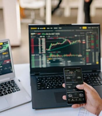 Auf einem Tisch befinden sich zwei Laptops und ein Smartphone. Die Bildschirme zeigen Finanzdaten und Diagramme. Diese technische Ausstattung könnte im Kontext der neuen White-Label-Lösung der Sutor Bank für das Altersvorsorge-Depot genutzt werden.