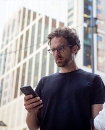 Ein Mann mit lockigem Haar und Brille betrachtet konzentriert sein Smartphone. Die moderne Architektur im Hintergrund deutet auf ein urbanes Umfeld hin. Jonas F. Sowa, Gründer von Digital8, könnte in diesem Kontext für digitale Sichtbarkeit stehen.