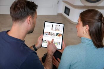 Zwei Personen betrachten gemeinsam auf einem Tablet Inhalte, die möglicherweise mit Immobilien in Verbindung stehen. Der Raum wirkt modern und hell, was auf eine zeitgemäße Wohnatmosphäre hinweist.