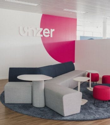Der Raum präsentiert eine moderne Lounge mit einem großen, blauen Sofa und mehreren Tischen. Rote und graue Sitzgelegenheiten ergänzen die Einrichtung. An der Wand ist das Logo von Unzer in weißer Schrift auf pinkem Hintergrund angebracht.