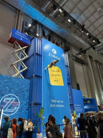 Der Messestand von Zurich auf dem OMR-Festival 2026 präsentiert sich eindrucksvoll mit blauen Containern. Eine Person klettert an der Wand, während Besucher anstehen. Slogans betonen die Kundenorientierung der Marke.