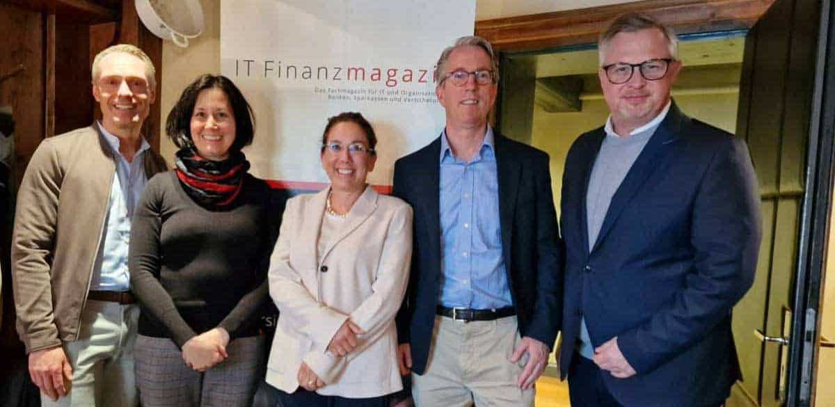 Fünf Personen stehen vor einem Banner des IT Finanzmagazins. Die Gruppe diskutiert über aktuelle Entwicklungen im Bereich Karten und deren Einfluss auf die Bankenbranche. Die Atmosphäre wirkt professionell und fokussiert.