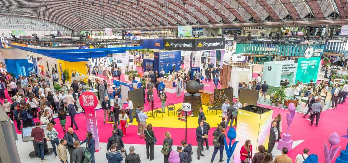 Die Veranstaltung Money20/20 präsentiert sich mit zahlreichen Ständen und Besuchern in einer großen Halle. Verschiedene Unternehmen, darunter Visa und Paymentwall, sind vertreten. Die Atmosphäre ist geschäftig, mit einem bunten Bodenbelag und vielfältigen Ausstellungsständen.