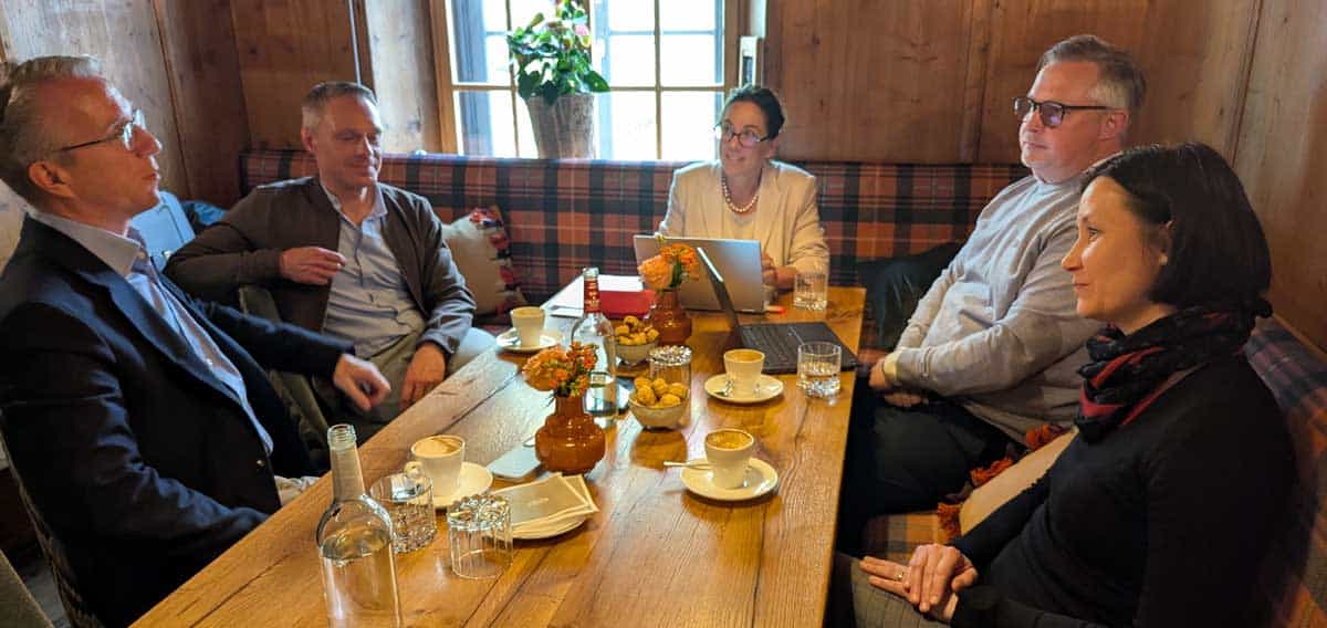 In einem rustikalen Raum diskutieren fünf Personen an einem Holztisch über Finanzthemen. Auf dem Tisch stehen Getränke und Snacks. Die Atmosphäre ist informell, was den Austausch über Karten und Finanzbildung fördert.