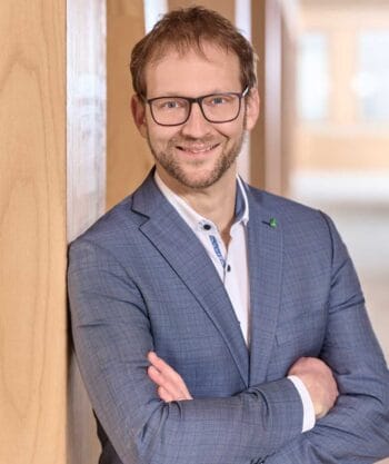 Dietmar von Blücher, Vorstandssprecher der UmweltBank, präsentiert sich in einem professionellen Umfeld. Er trägt einen blauen Anzug und lächelt, während er mit verschränkten Armen vor einer Holzverkleidung steht. Der Hintergrund ist hell und modern gestaltet.