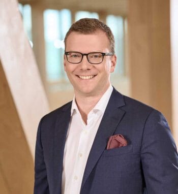 Benjamin Schmidt, IT-Leiter UmweltBank, präsentiert sich in einem professionellen Umfeld. Er trägt einen Anzug und lächelt. Im Hintergrund sind helle, moderne Architekturmerkmale zu erkennen, die eine innovative Atmosphäre vermitteln.