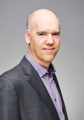 Michael Pattison, Principal Solutions Engineer bei Okta, präsentiert sich in einem grauen Anzug mit einem karierten Hemd. Sein Blick ist direkt und neutral. Die Darstellung vermittelt Professionalität und Expertise im Bereich DPoP und Sicherheitsarchitektur.