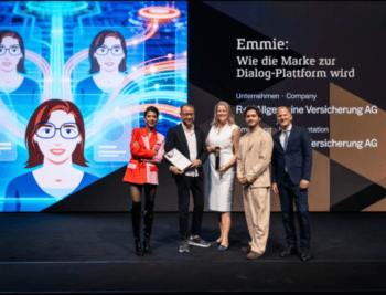 Auf der Bühne präsentieren mehrere Personen die Auszeichnung für den KI-Chatbot Emmie. Im Hintergrund ist eine digitale Darstellung von Emmie zu sehen. Die Atmosphäre ist feierlich, und die Auszeichnung wird für die Kategorie „Best AI Project of the Year“ überreicht.