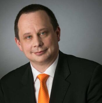 Ein Mann in einem schwarzen Anzug mit orangefarbener Krawatte blickt direkt in die Kamera. Die Hintergrundfarbe ist neutral. In der Diskussion um Core-Banking-Fraud-Systeme wird die Relevanz biometrischer Daten und Device-Fingerprints thematisiert.