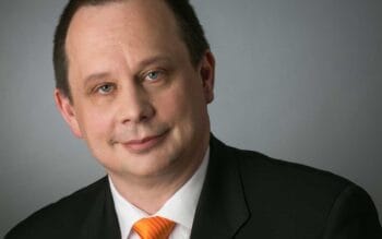 Frank Meltke, CEO von Contraco, präsentiert sich in formeller Kleidung mit einem orangefarbenen Krawatte. Seine Expertise in digitalen Transformationsberatungen und Core-Banking-Fraud-Systemen ist durch seine langjährige Erfahrung und Patente belegt.