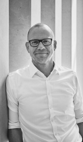 Christian von Hammel-Bonten, ein Experte im Bereich Digitaler Euro, präsentiert sich in einem formellen Hemd vor einer minimalistischen Architektur. Sein Lächeln und die entspannte Körperhaltung vermitteln Offenheit und Zugänglichkeit in der Diskussion über digitale Zahlungsmethoden.