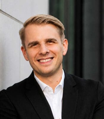 Ein lächelnder Mann in einem schwarzen Anzug steht vor einer modernen Architektur. Die Agent Pay Transaktion von Mastercard in Deutschland wird als bedeutender Fortschritt im Agentic Commerce betrachtet. Details zur Person sind nicht verfügbar.