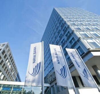 Die Aufnahme zeigt die Fassade des Gebäudes der NRW.BANK, umgeben von mehreren Fahnen mit dem Logo der Bank. Der Himmel ist klar und blau, was eine moderne und einladende Atmosphäre vermittelt.