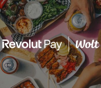 Im Bild sind verschiedene Speisen in Schalen und auf Tellern angeordnet, während Hände eine Dose und eine Schale halten. Das Logo von Revolut Pay ist prominent platziert, was die Integration in die Wolt-Plattform verdeutlicht.