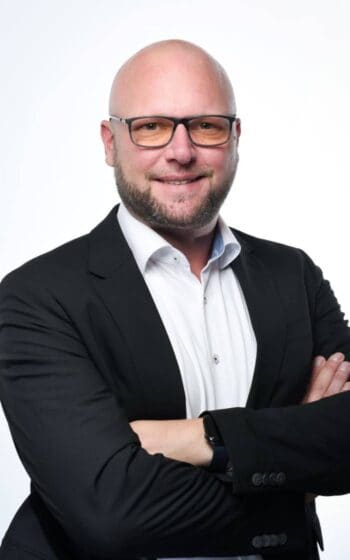 Sebastian Kolter, Senior Director Banking bei Diebold Nixdorf, präsentiert sich in einem formellen Outfit mit verschränkten Armen. Der Hintergrund ist neutral, was die Professionalität im Bereich Bargeldmanagement unterstreicht.